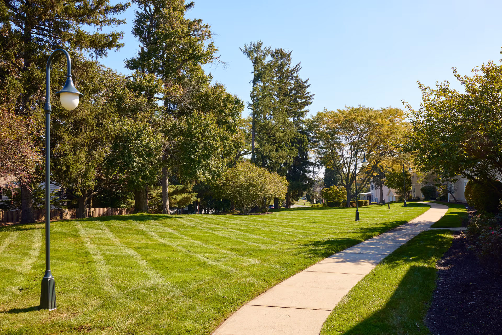 A sunny outdoor scene at Brandywine Governor's Crossing by Monarch featuring a well-maintained grassy area with a paved walking path curving through it. There are several tall trees and lamp posts lining the path, with some buildings partially visible in the background under a clear blue sky.