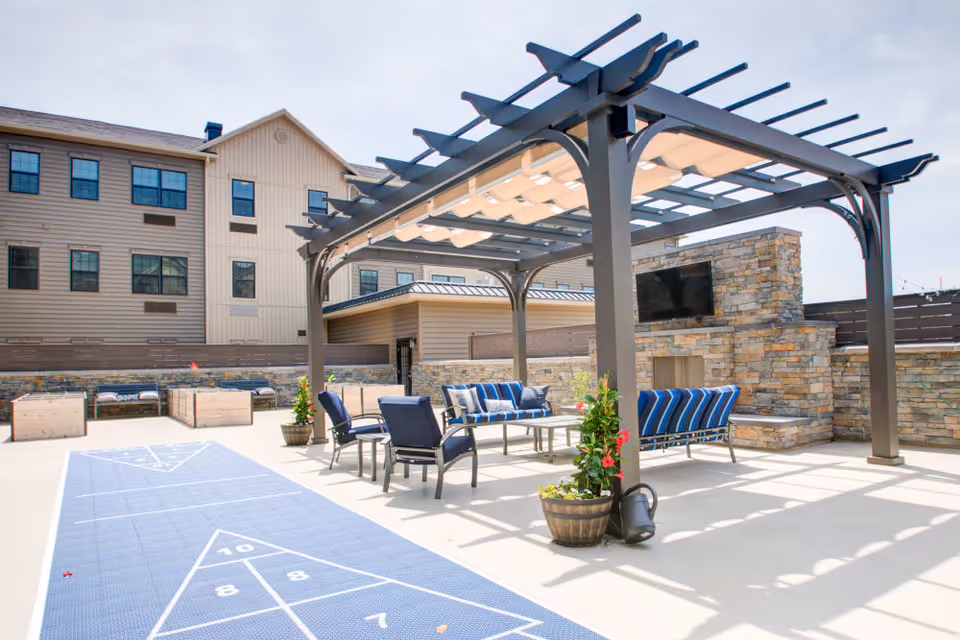 Outdoor patio area at The Pinnacle at Plymouth Meeting featuring a pergola with seating underneath, including cushioned chairs and sofas. There is a stone fireplace with a mounted TV on the right side. The area also includes a shuffleboard court in the foreground and planter pots with flowers.