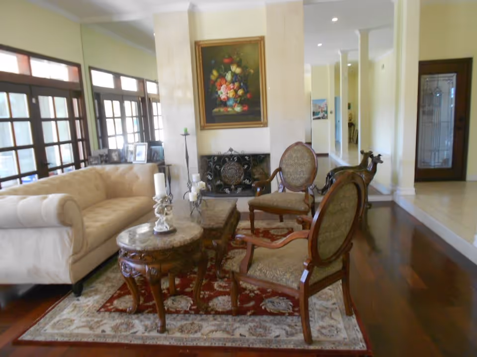 A cozy living room with a beige tufted sofa, two upholstered wooden armchairs, a round wooden coffee table with candles, and a rectangular wooden table on a patterned area rug. The room has large windows with wooden frames, a fireplace with an ornate screen, and a floral painting above it. The floor is dark wood, and the walls are light-colored.