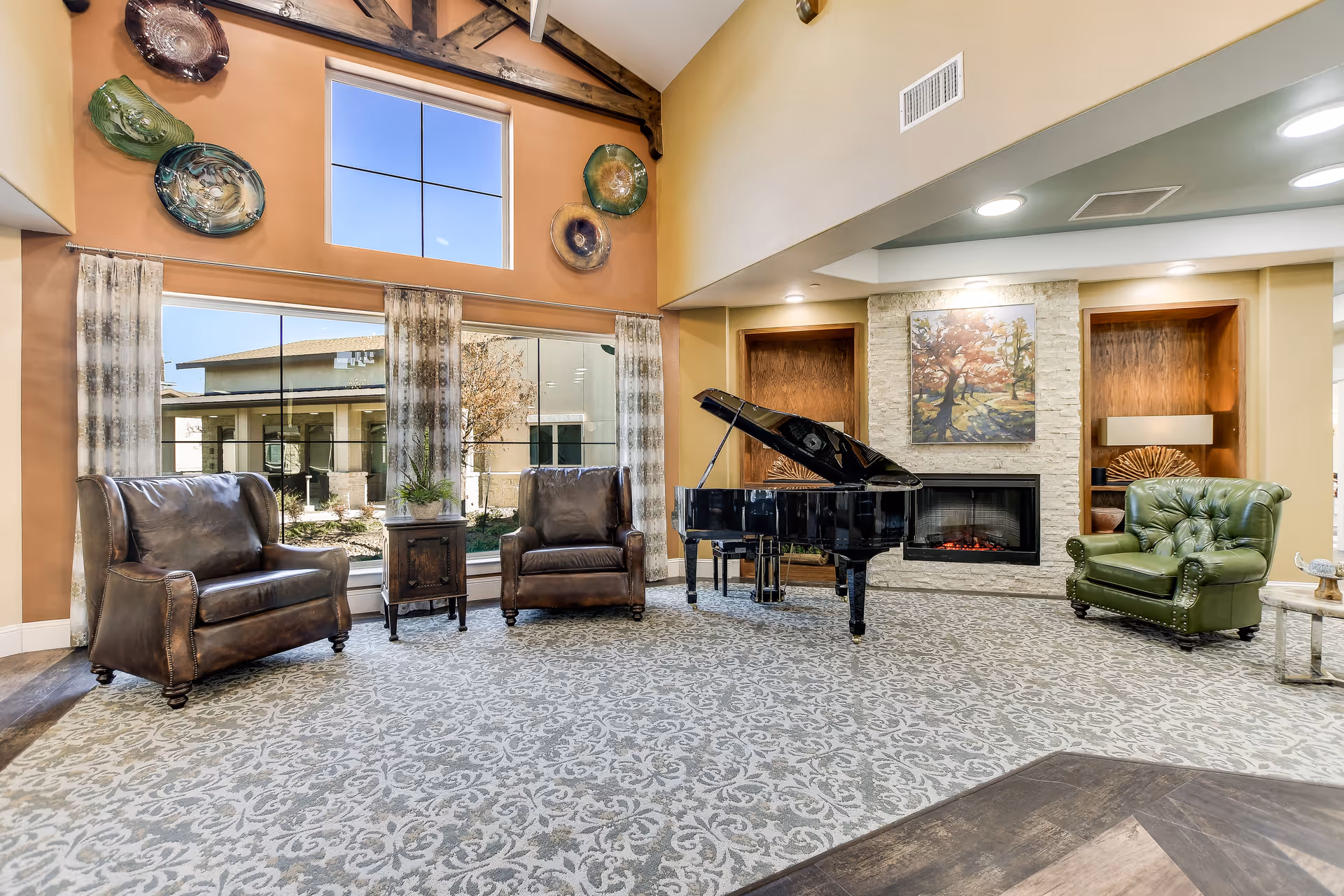 A spacious senior living facility common area with high ceilings and large windows. The room features two brown leather armchairs, one green leather armchair, a black grand piano, and a fireplace with a painting of a tree above it. Decorative glass wall art is mounted on the wall above the windows, and patterned carpet covers the floor.