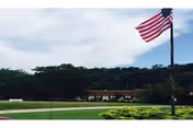 Flagpole with waving American flag in front of grassy area and distant building.