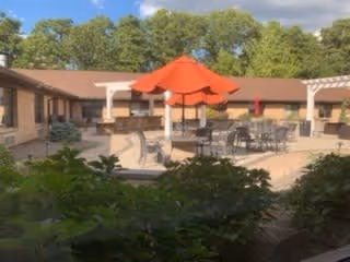 Outdoor courtyard area at Harmony Village at CareOne Jackson featuring patio tables with umbrellas, surrounded by a single-story building and greenery under a partly cloudy sky.