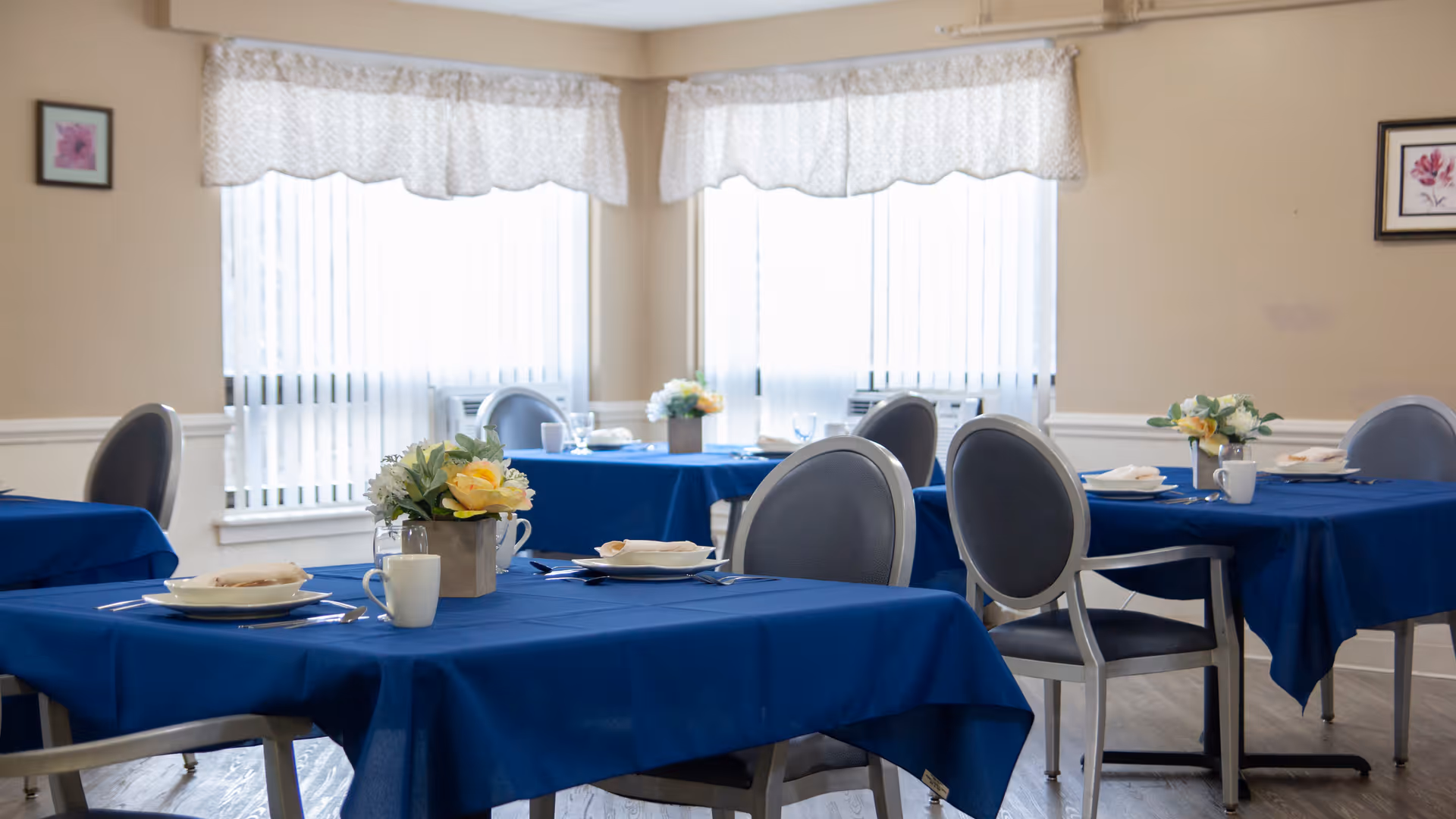 A dining room with several tables covered in blue tablecloths, each set with plates, cups, and silverware. The tables have small flower arrangements as centerpieces. The room has beige walls, large windows with white curtains, and framed floral artwork on the walls.
