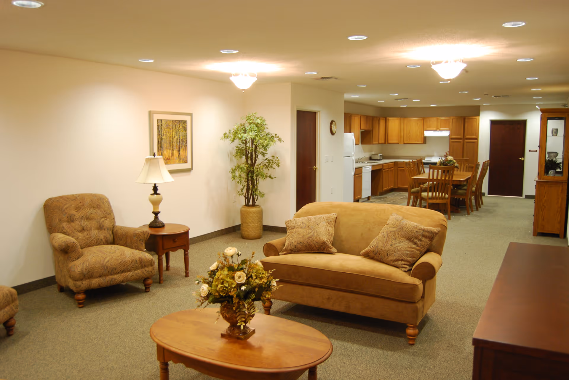 A cozy senior living common area featuring a beige loveseat with two patterned cushions, a matching armchair, a wooden coffee table with a floral arrangement, and a side table with a lamp. In the background, there is a kitchen with wooden cabinets, a dining table with chairs, and a potted plant near the wall. The room is well-lit with ceiling lights and has a carpeted floor.