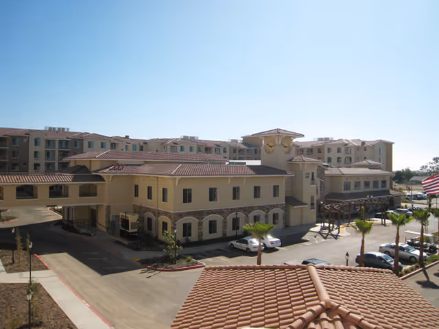 Exterior view of a senior living facility with multiple connected buildings featuring beige walls and red-tiled roofs. The main building has a clock tower, and there are several palm trees and parked cars in front. The sky is clear and blue.