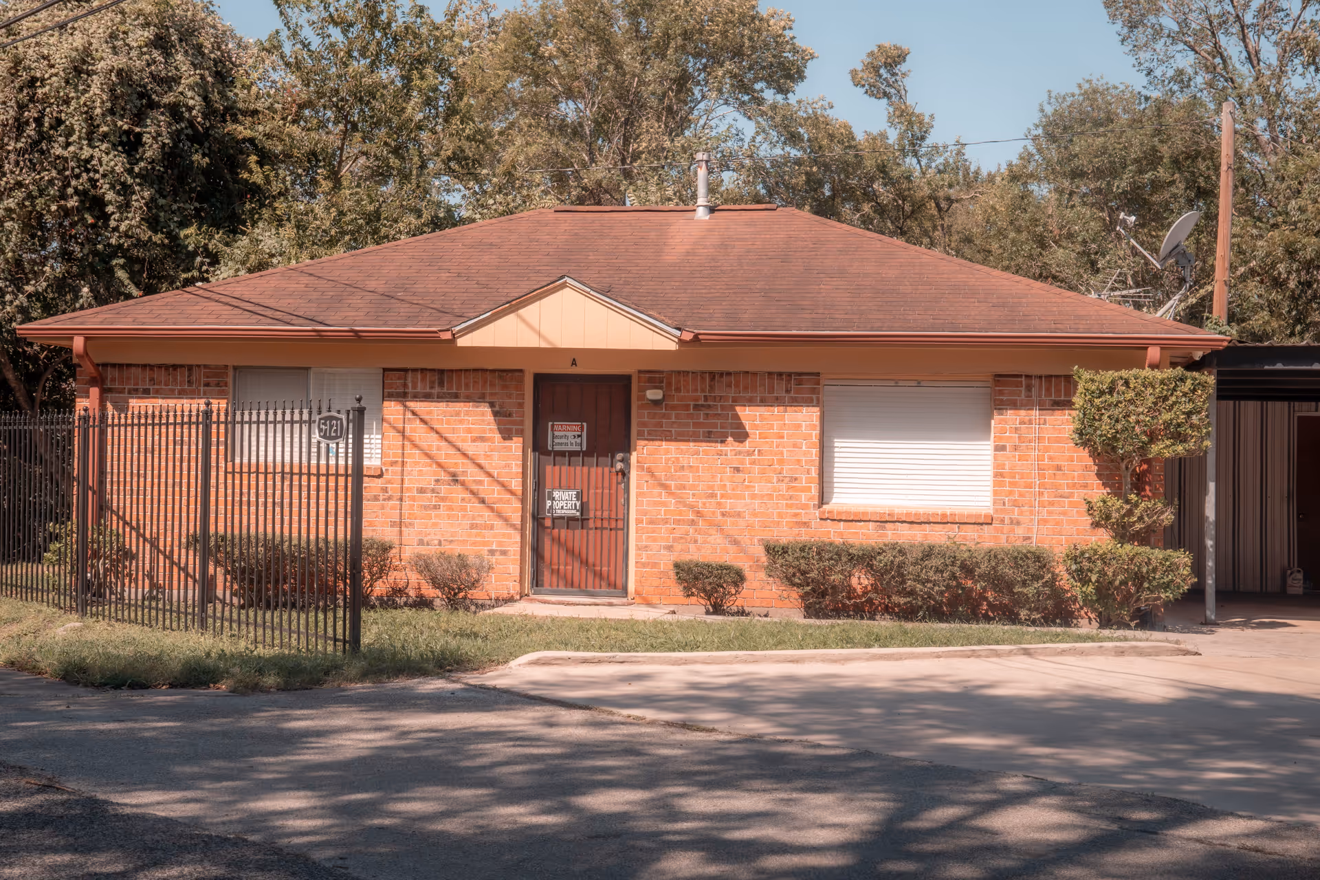 Single-story brick building with a brown shingled roof, a metal fence on the left side, and trimmed bushes in front. The building has a door with signs and two windows with closed blinds. Trees are visible in the background under a clear sky.