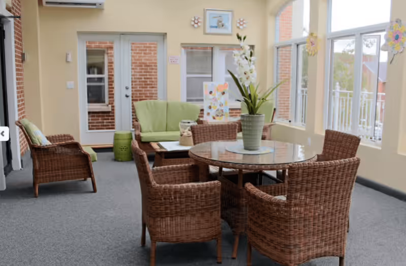 A bright indoor seating area with wicker furniture including a round glass-top table surrounded by four wicker chairs, a green cushioned loveseat, and a wicker armchair. There is a potted plant with white flowers on the table, large windows letting in natural light, and a door leading outside. The walls are light yellow and decorated with colorful flower-shaped wall hangings.