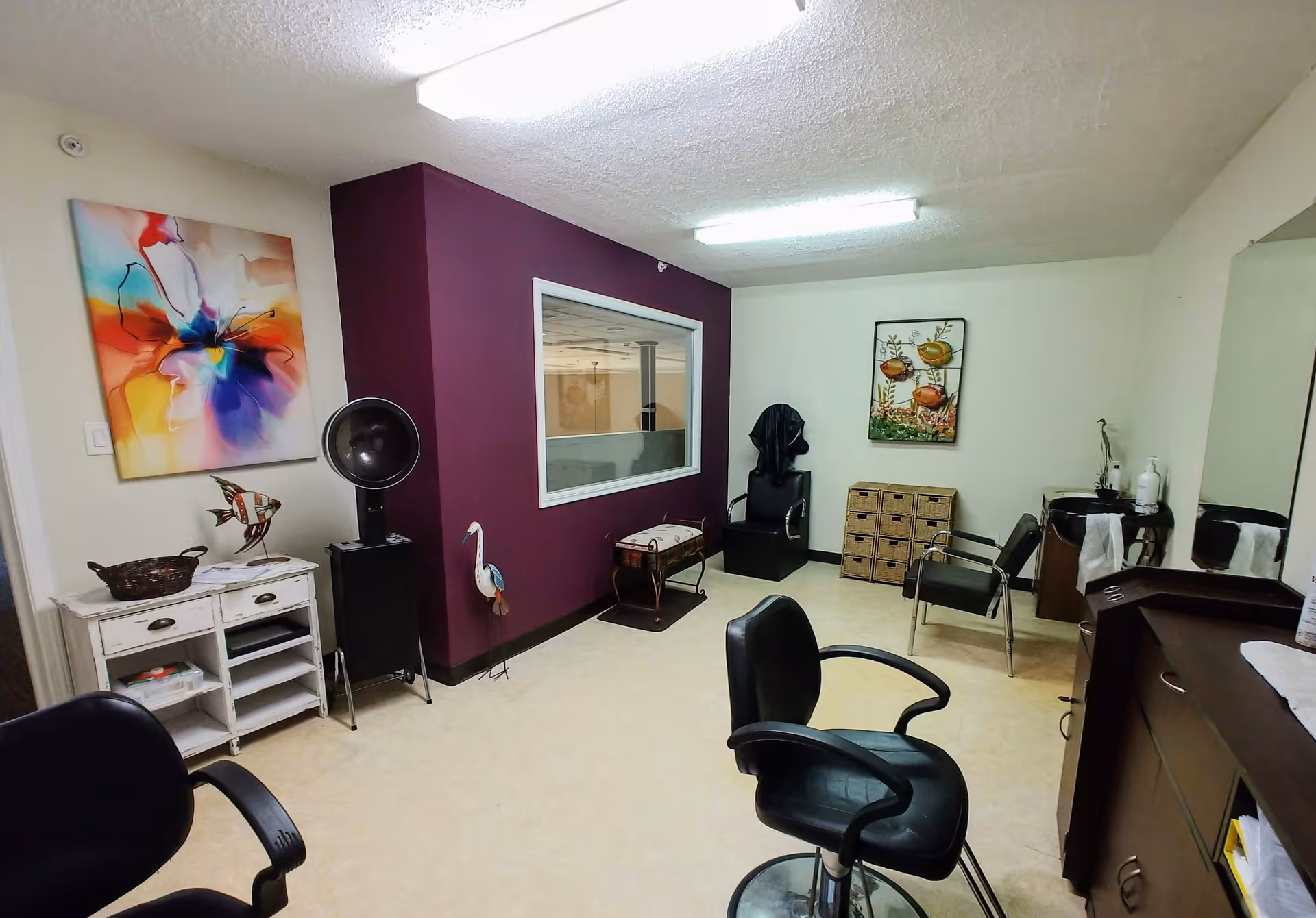 Interior of a salon room with several black salon chairs, a hair dryer, a small white cabinet, colorful wall art, a purple accent wall with a window, and a wicker drawer unit. The room is well-lit with fluorescent lights and has a clean, organized appearance.