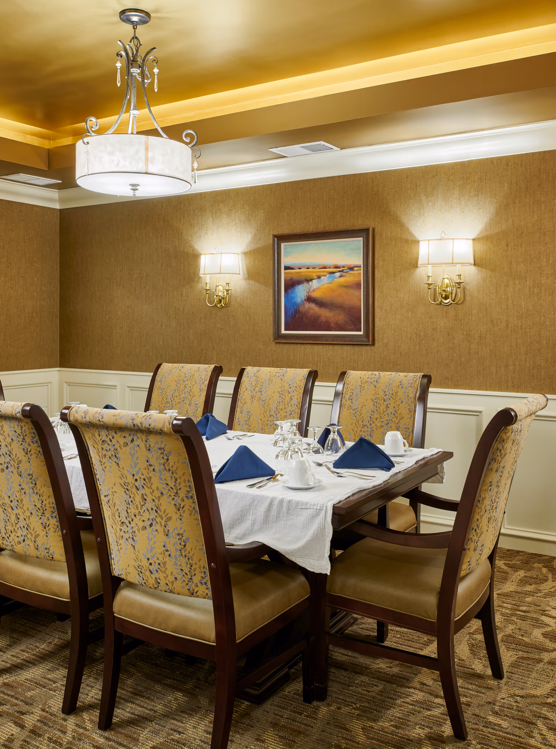 A formal dining room with a set table, upholstered chairs, a chandelier, wall sconces, and a landscape painting on the wall.