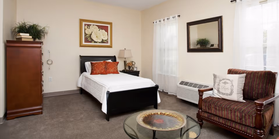 A cozy bedroom with a single bed dressed in white bedding and two red decorative pillows. To the left of the bed is a wooden dresser topped with a plant and books. A nightstand with a lamp and clock sits next to the bed. On the right side, there is a striped armchair with a decorative pillow and a glass coffee table with a decorative plate. The room has beige walls, a framed floral painting above the bed, a large mirror on the wall, and two windows with white curtains.