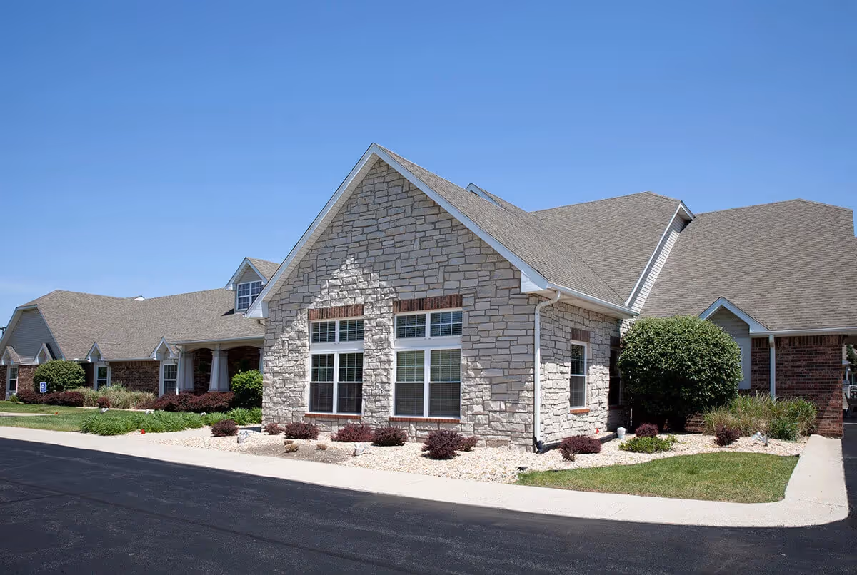 Front exterior of a single-story stone and brick senior living building with landscaped beds and a driveway.
