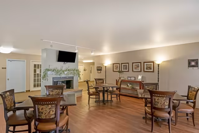 Spacious senior living common room with multiple round tables and chairs, a fireplace with a wall-mounted TV, and framed artwork on the walls.