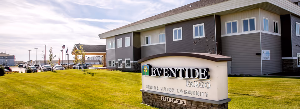 Exterior view of the Eventide Fargo senior living community building with a large sign in the foreground displaying the facility name and address. The building is two stories with multiple windows, surrounded by a well-maintained lawn and a parking lot with several cars. The sky is partly cloudy.