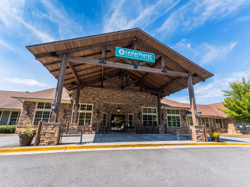 Front exterior view of Cedarhurst Senior Living of Canton building with a large covered entrance supported by wooden beams, stone facade, and multiple windows under a blue sky with some clouds.