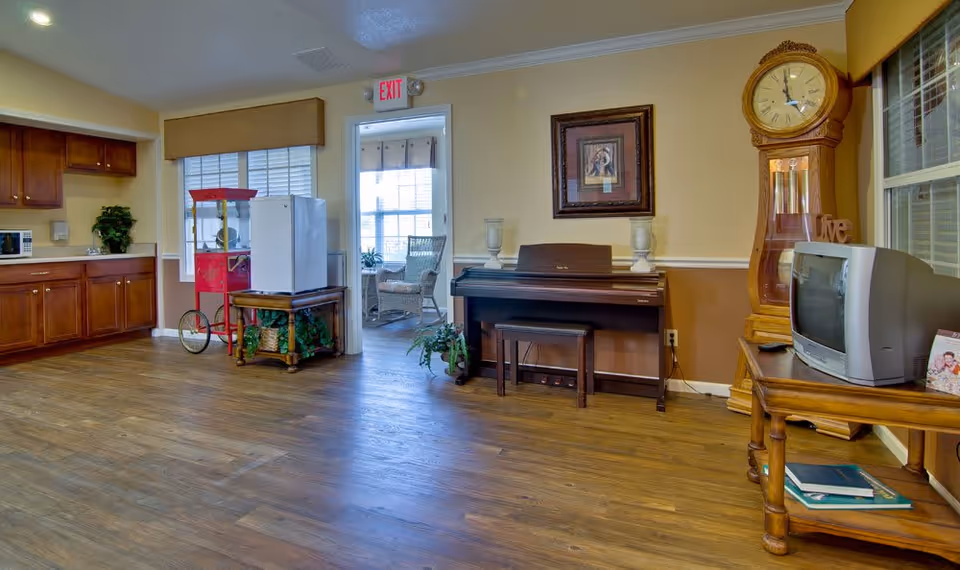 Community common room with wooden floors, a piano, grandfather clock, TV on a table, kitchenette cabinets, and a popcorn machine.