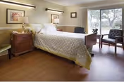 A well-lit bedroom in a senior living facility featuring a neatly made bed with white bedding, a wooden nightstand with a lamp and framed artwork on the wall. There are two upholstered chairs near a large window that offers a view of trees outside.