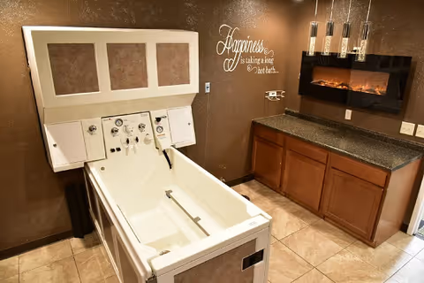 A bathing room with a specialized assisted living bathtub featuring control knobs and handrails. The room has brown walls, a countertop with wooden cabinets, and a wall-mounted electric fireplace. A decorative wall decal reads, 'Happiness is taking a long hot bath...'.