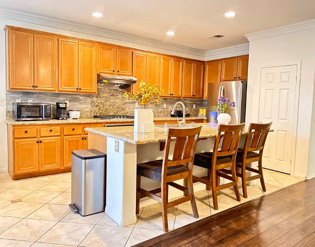 A bright kitchen with wooden cabinets, granite countertops, and a kitchen island with three wooden chairs. The kitchen features a stainless steel refrigerator, microwave, coffee maker, and a trash bin. There are decorative plants on the island and recessed lighting on the ceiling.
