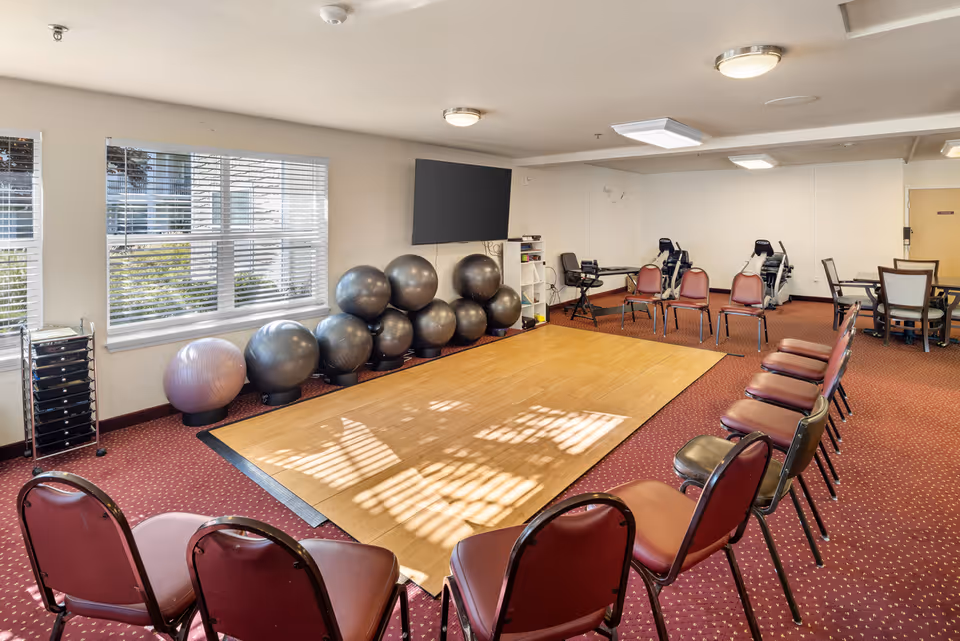 A senior living facility activity room with a wooden floor exercise area surrounded by chairs. There are several large exercise balls stacked against the wall under a mounted flat screen TV. The room has windows with blinds letting in natural light, exercise equipment in the background, and a table with chairs in the corner.