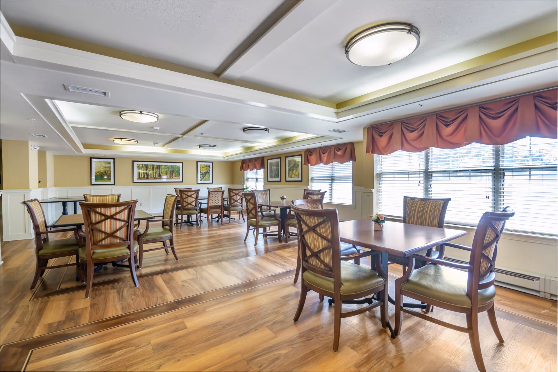 Bright dining room with wooden tables and chairs, large windows with valances, and framed artwork on the walls.