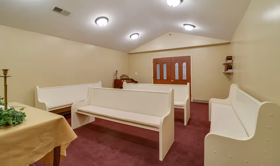 Interior of a small chapel or meditation room with cream-colored walls and ceiling, maroon carpet, white wooden pews arranged in rows, a table with a beige cloth and a candle holder with greenery, and double wooden doors with glass panels at the far end.
