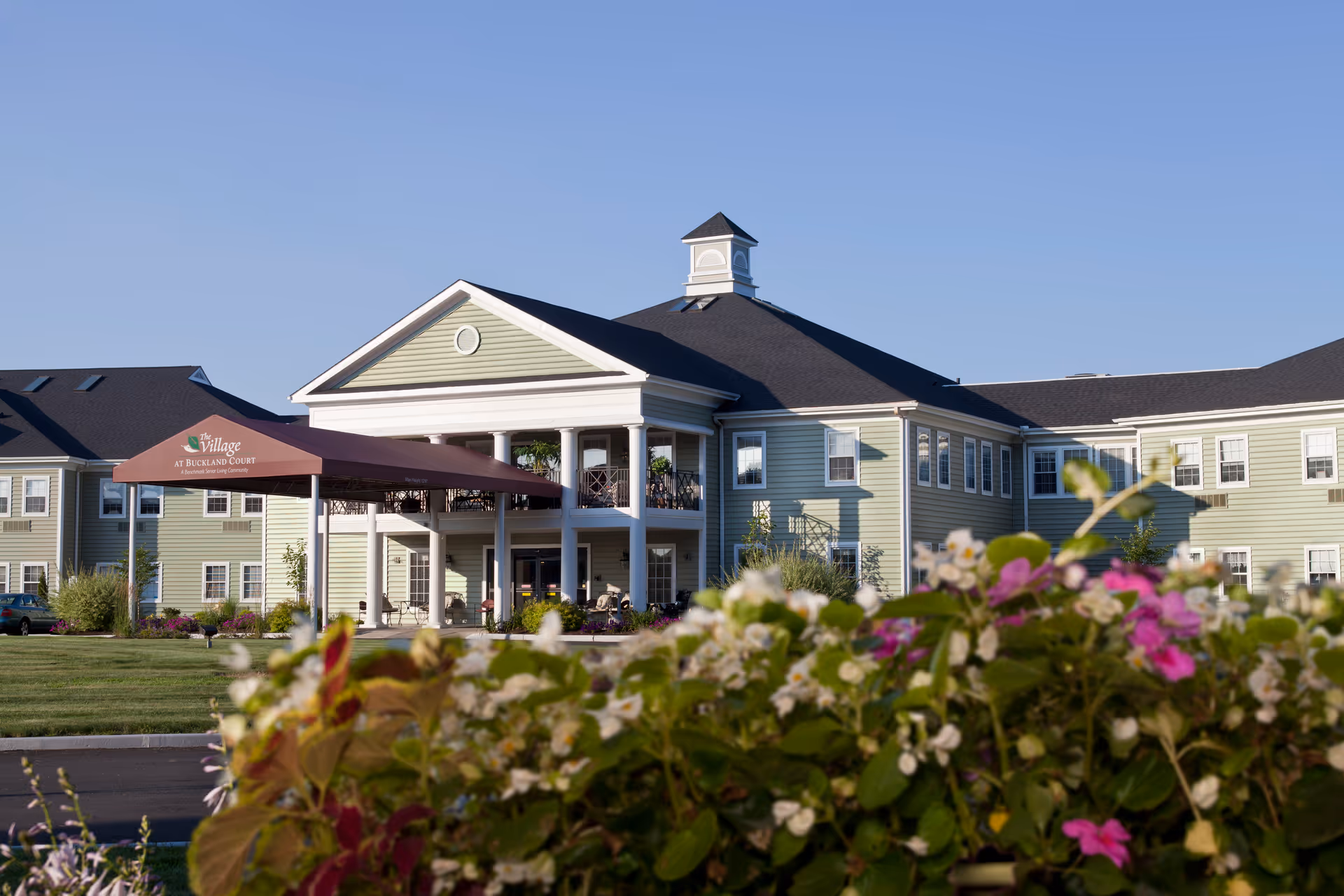 Exterior view of The Village at Buckland Court senior living facility with a large entrance canopy, green siding, white trim, and a cupola on the roof. Flowers and greenery are in the foreground under a clear blue sky.