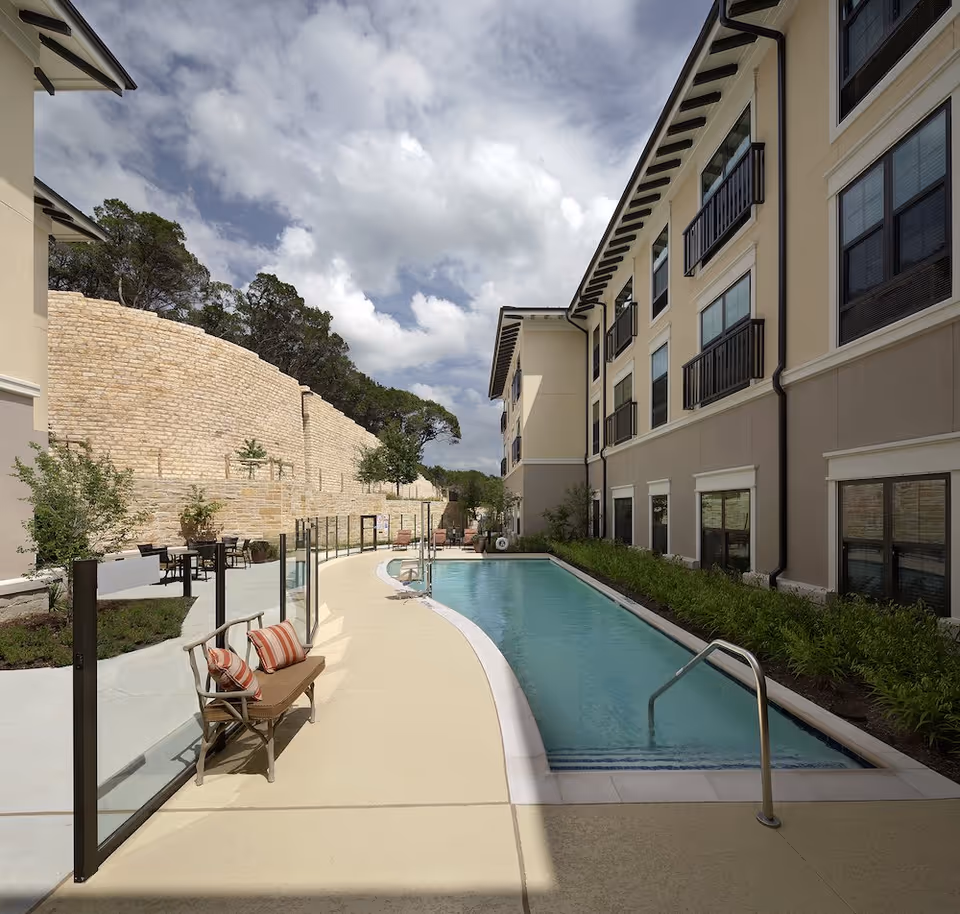 Outdoor courtyard with a narrow swimming pool alongside a multi-story senior living building, a bench with cushions and glass safety fencing under a partly cloudy sky.