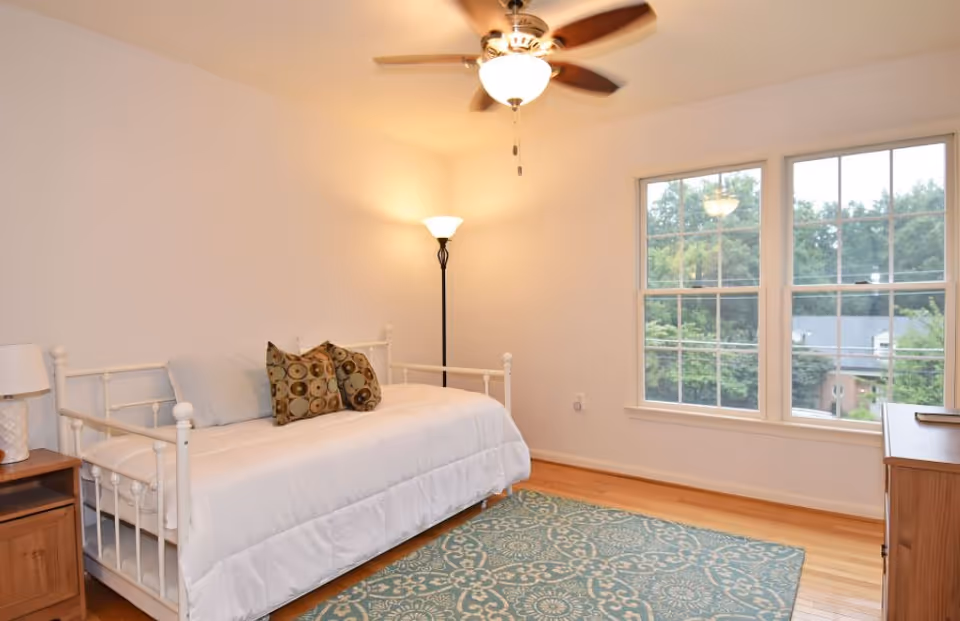 A bright bedroom with a white metal daybed covered with a white comforter and two patterned pillows. There is a wooden nightstand with a lamp on the left side of the bed, a tall floor lamp in the corner, a ceiling fan with a light, and a large window showing green trees outside. The floor is wooden with a blue patterned area rug.
