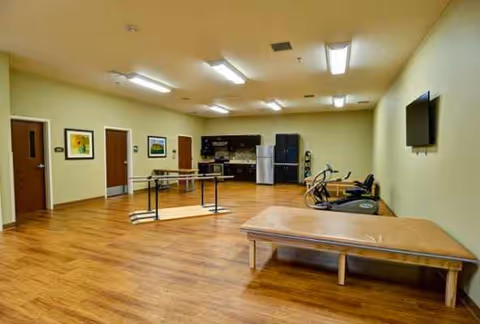 A spacious senior living activity room with wooden floors, tables, exercise equipment, and a wall-mounted TV.