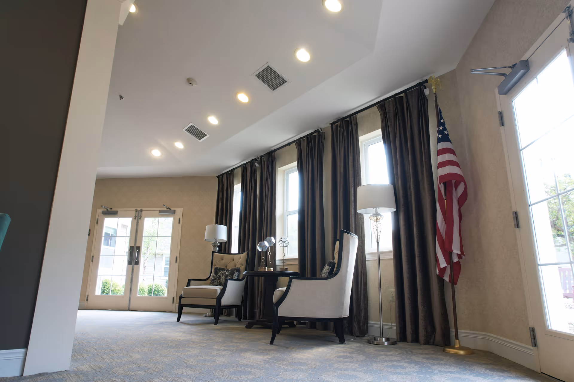 A bright and spacious seating area in a senior living facility with two upholstered armchairs, a small round table with decorative items, two floor lamps, large windows with dark curtains, and an American flag in the corner near glass doors leading outside.