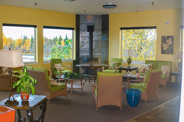 A cozy living room area with yellow walls and large windows showing trees outside. The room features green cushioned wicker chairs arranged around a central stone fireplace with a wood stove. There are side tables with lamps and decorative plants, creating a warm and inviting atmosphere.