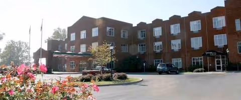 Brick nursing facility with multiple windows, a parking lot, and flowering foreground.