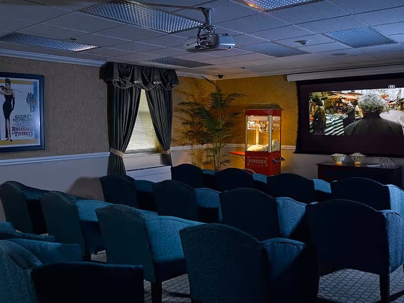 A small movie theater room with rows of blue upholstered chairs facing a large screen showing a movie scene. There is a popcorn machine near the screen, a window with dark curtains, a potted plant, and a framed Audrey Hepburn poster on the wall.
