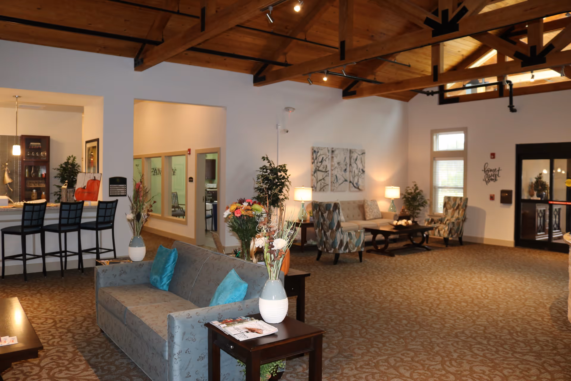 A spacious and well-lit living room area in a senior living facility with a high wooden beam ceiling. The room features a gray sofa with blue cushions, two patterned armchairs, a coffee table, side tables with lamps, and decorative plants. There is a counter with three black bar stools on the left side and a hallway leading to other rooms. The carpet has a subtle pattern, and the walls are painted white. A glass door is visible on the right side.
