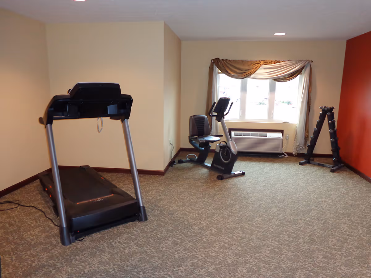 A small exercise room with a treadmill, a recumbent stationary bike, and a rack of dumbbells. The room has beige walls with one red accent wall, a window with curtains, and carpeted flooring.
