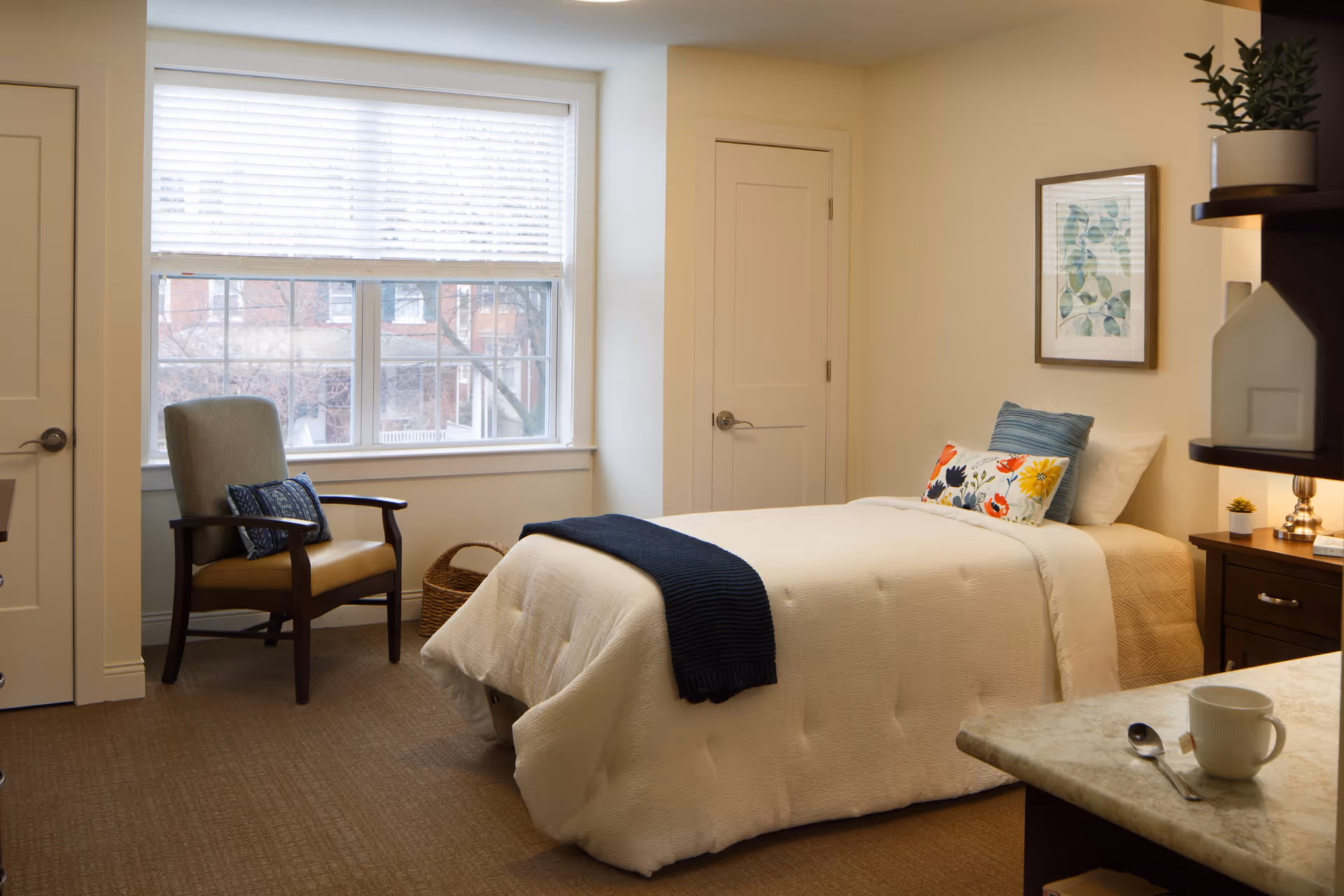 A cozy bedroom with a single bed covered in a white quilt and a dark blue throw blanket. The bed has two decorative pillows, one blue and one floral. There is a wooden nightstand with a lamp and small plant, a framed botanical print on the wall, a cushioned armchair with a blue pillow near a window with white blinds, and a basket on the floor. A countertop with a white cup, spoon, and tea bag is visible in the foreground.