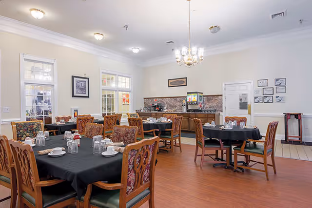 A dining room with several wooden tables covered with black tablecloths, set with cups, glasses, and napkins. The room has wooden chairs with patterned cushions, a beverage dispenser on a counter against the far wall, and framed pictures on the walls. The floor is wooden, and the ceiling has a chandelier and recessed lighting.