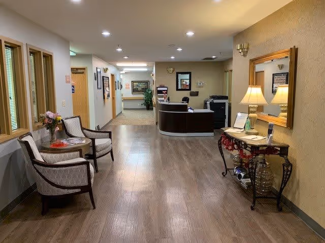 A well-lit hallway in a senior living facility with wooden flooring and beige walls. On the left side, there are two upholstered chairs with a small round table holding a flower vase. On the right side, there is a decorative console table with a lamp, a mirror above it, and some decorative items underneath. At the end of the hallway is a reception desk with office equipment and a plant in the background.
