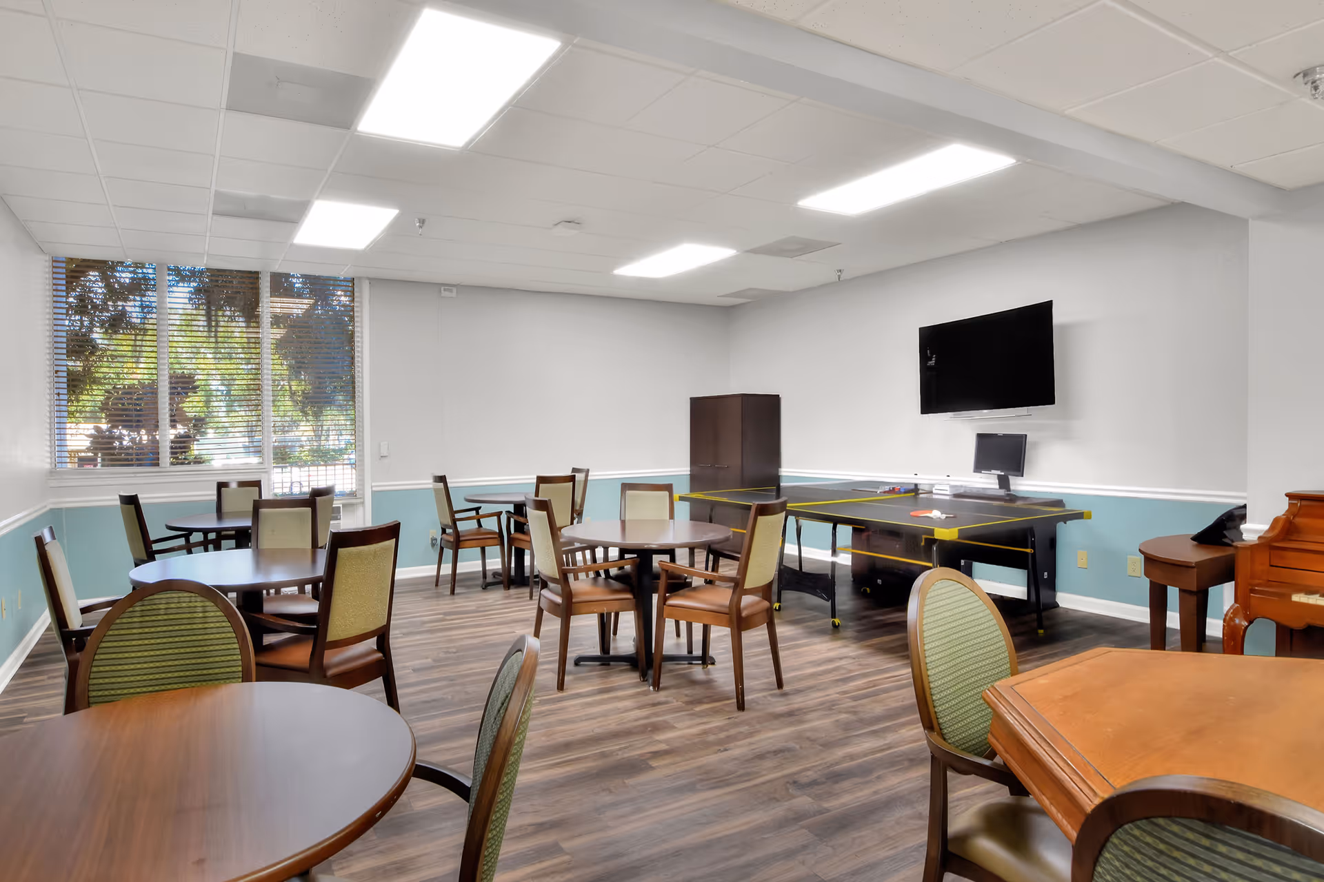 A well-lit common room with several round tables and chairs arranged around the room. There is a large window with blinds on the left wall, a flat-screen TV mounted on the right wall above a table, and a piano in the corner. The room has wood flooring and light blue and white walls.