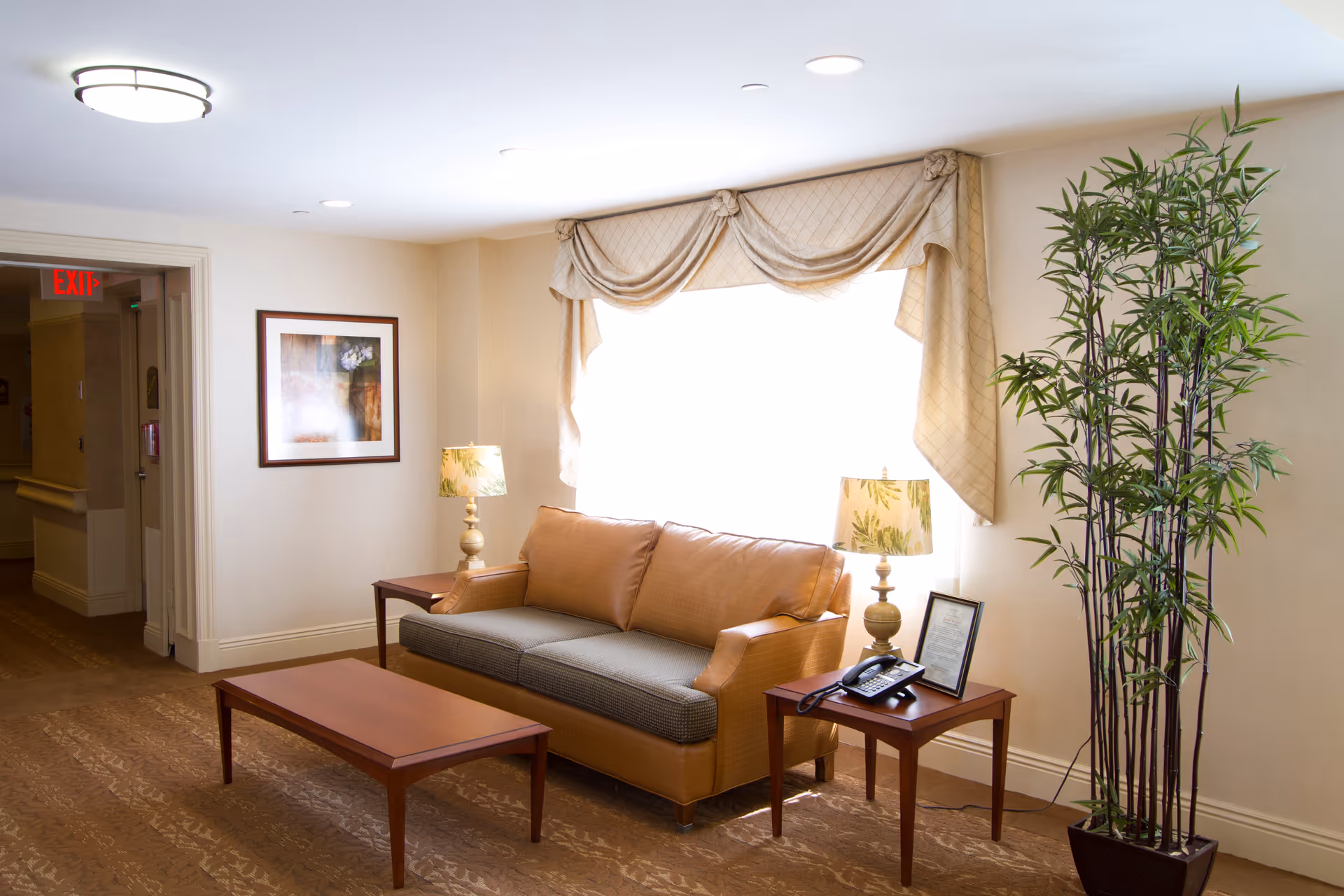 A cozy seating area in a senior living facility with a tan sofa, two wooden side tables each holding a lamp with a leafy pattern, a wooden coffee table, a framed picture on the wall, a large window with beige drapes, and a tall potted plant in the corner.