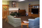 A cozy living room area with two upholstered armchairs and a wooden chair arranged around a small wooden coffee table. There is a fireplace with a framed picture above it, a piano against the wall, and a wooden entertainment center with a television and decorative items. The room has carpeted flooring and neutral-colored walls.