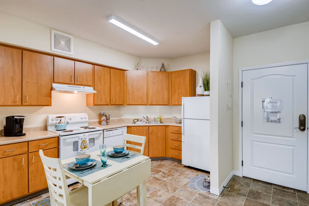 A kitchen area with wooden cabinets, a white stove, dishwasher, and refrigerator. A small dining table set for two with blue dishes and glasses is in the foreground. The floor is tiled, and there is a white door on the right side of the image.