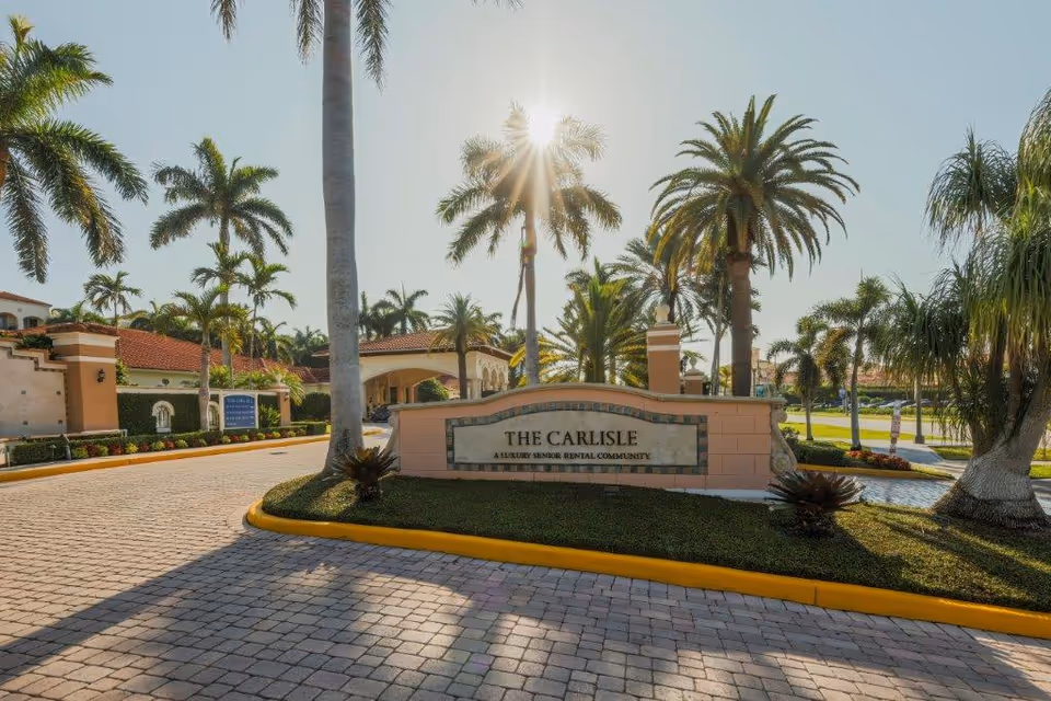 Entrance sign for The Carlisle, a luxury senior rental community, surrounded by palm trees and landscaped greenery with a paved driveway under a clear sky with the sun shining through the palm leaves.