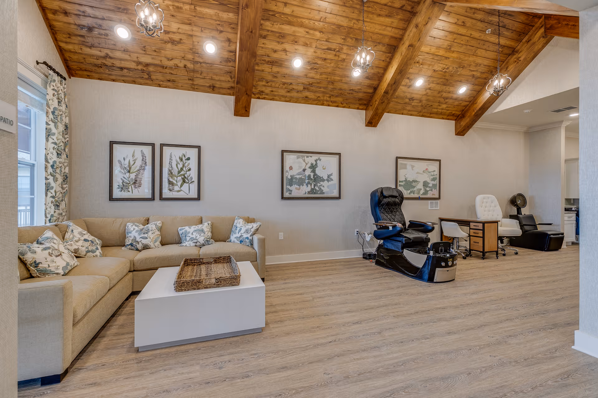 A spacious room with a wooden vaulted ceiling featuring exposed beams and pendant lights. The room has a beige sectional sofa with floral patterned cushions, a white coffee table with a wicker tray, and three framed botanical prints on the wall. On the right side, there are two pedicure chairs and a small desk with a white office chair and a fan. The floor is light wood, and a window with floral curtains is visible on the left.