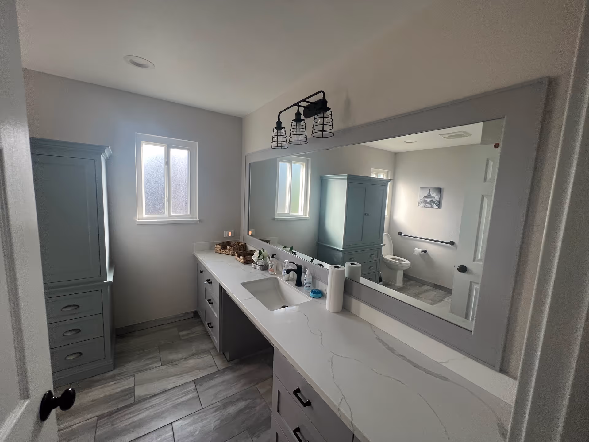 Accessible bathroom with a long white vanity and sink, large mirror, storage cabinets, tiled floor, and a toilet with a grab bar.