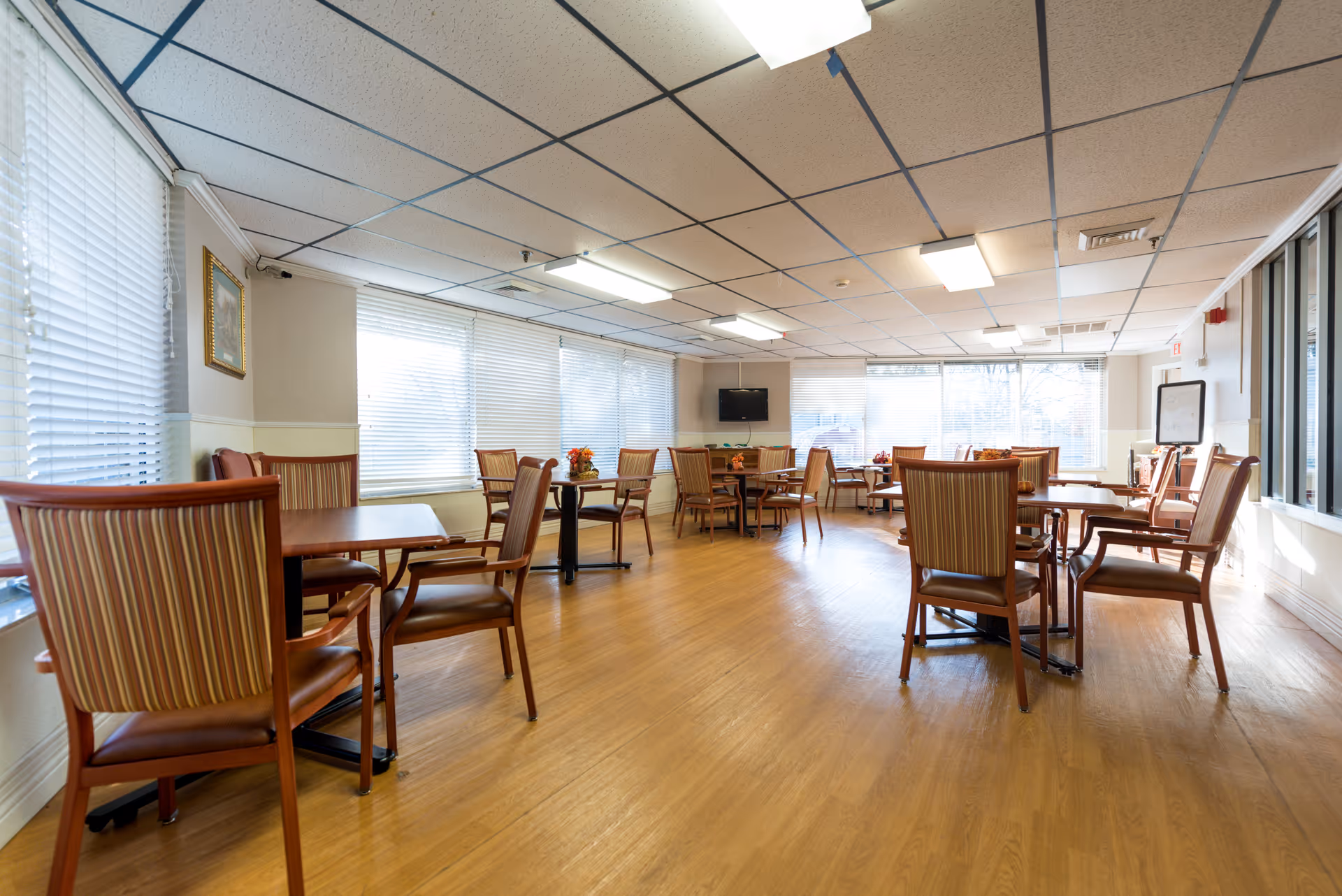 A bright dining room with multiple wooden tables and striped cushioned chairs arranged neatly. Large windows with blinds allow natural light to fill the room. A flat-screen TV is mounted on the far wall, and small flower arrangements are placed on the tables. The floor is wooden, and the ceiling has fluorescent lighting panels.
