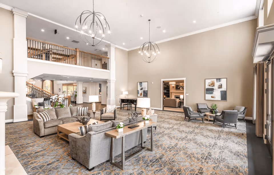Spacious senior living facility common area with high ceilings, modern chandeliers, multiple seating arrangements including sofas and armchairs, a grand piano, and decorative artwork on the walls. The room features a patterned carpet and large windows allowing natural light.