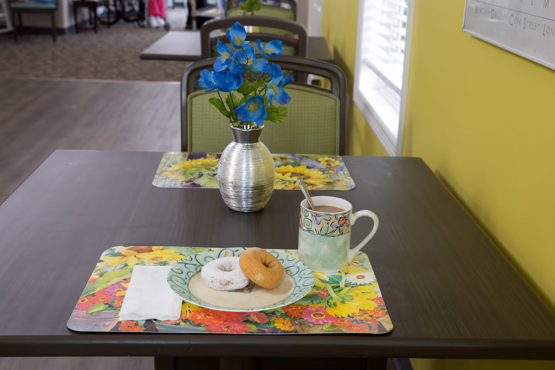 A dining table set with a floral placemat, a plate holding two donuts, a napkin, and a mug with a spoon. A silver vase with blue flowers is placed on the table. The background shows more tables and chairs in a well-lit room with yellow walls and a window with blinds.