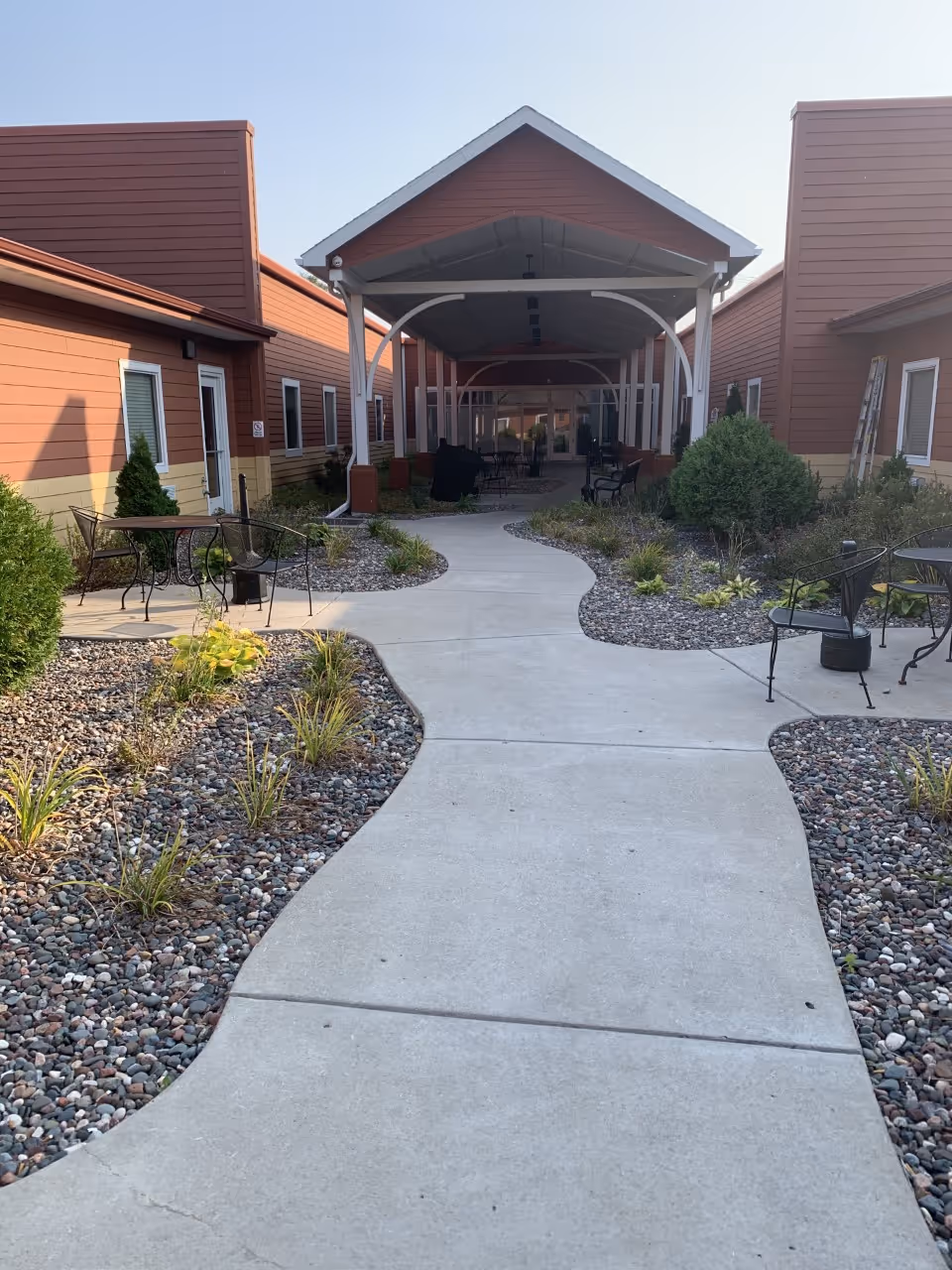 Outdoor courtyard area with a winding concrete pathway flanked by small plants and rocks. There are metal chairs and tables on either side of the path. The courtyard is surrounded by red and beige buildings with windows and doors, and a covered walkway with white pillars extends through the center.