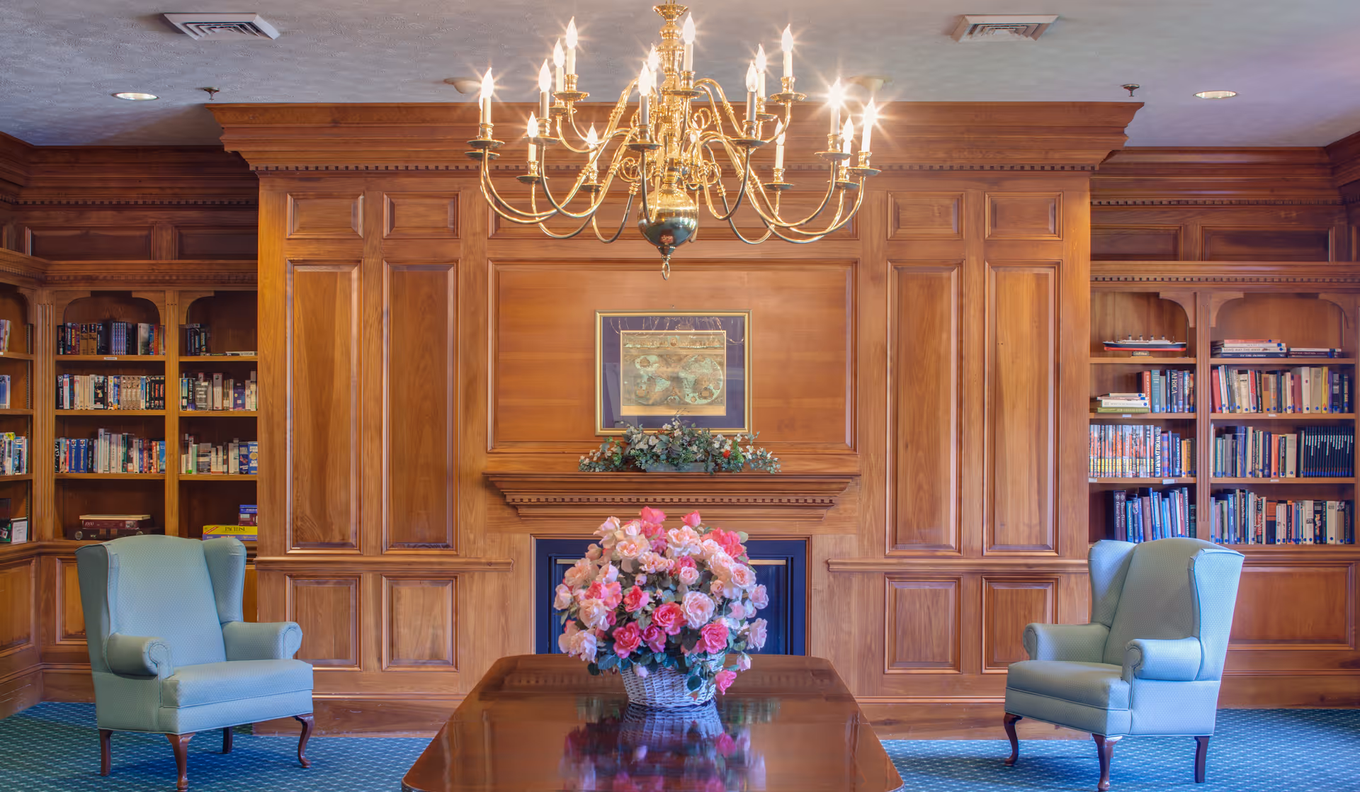 Elegant wood-paneled common room with bookshelves, a chandelier, two armchairs, and a table with a floral centerpiece.
